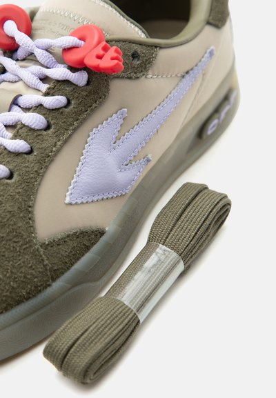 Μπεζ και ελιά αθλητικό παπούτσι με λογότυπο από δέρμα σε σχήμα βέλους σε λιλά χρώμα, μωβ κορδόνια με κόκκινα κλείστρα και ξεχωριστό τυλιγμένο κορδόνι ελιάς σε λευκό φόντο.