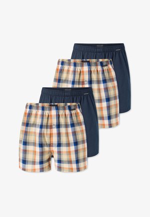 Uomo senza maglietta con capelli corti che indossa boxer a quadri arancioni, blu e beige, in piedi contro uno sfondo bianco.