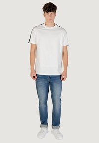 T-shirt bianca in cotone a maniche corte, con dettagli neri sulle spalle, abbinata a jeans in denim blu e sneaker bianche.