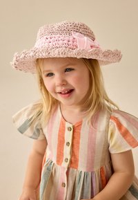 Chapeau de paille rose clair avec un ruban décoratif, doté d'un large bord et d'un tissage texturé. Enfant habillée d'une robe rayée multicolore.