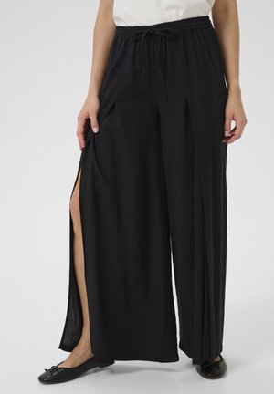 Frau in schwarzen weit geschnittenen Hosen mit seitlichen Schlitzern, kombiniert mit schwarzen Ballerinas, vor einem schlichten Hintergrund stehend.