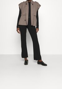 Bezramienna kamizelka w kolorze taupe z naramiennikami na ramionach, noszona na czarnym, prążkowanym topie oraz czarnych, rozszerzających się spodniach. Czarne loafersy dopełniają stylizację.