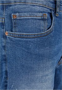 Vaqueros de mezclilla en azul claro con un bolsillo trasero visible. Presenta remaches de cobre, costuras naranjas contrastantes y una textura de lavado desvanecido.