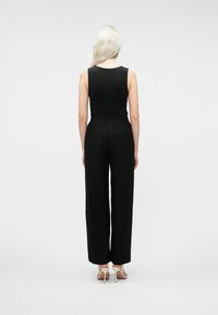 Blond kvinne vist fra ryggen iført en ermeløs svart topp, svarte høytaljede bukser og beige høyhælte sandaler.