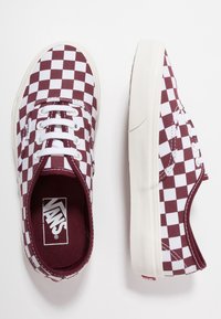 Paire de baskets Vans basse avec un motif damier bordeaux et blanc, lacets blancs et semelles épaisses en caoutchouc blanc sur fond blanc.