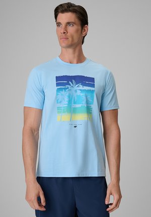 Mężczyzna w jasnoniebieskim t-shircie z grafiką palmy w odcieniach niebieskiego, zielonego i żółtego oraz w ciemnoniebieskich szortach, stojący na szarym tle.