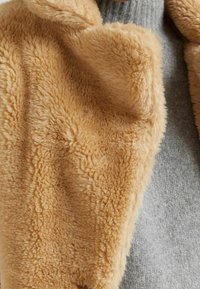 Chaqueta de pelaje sintético color beige con una superficie suave y texturizada, con un cuello drapeado y combinada con un suéter de punto gris debajo.