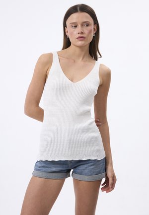 Valge sooniline tops, millel on V-kaelus ja laineline allserv. Kangas on tekstureeritud ning seda on kombineeritud lühikeste teksadega, mille allserv on üleskeeratud.