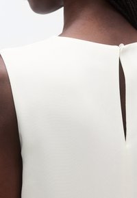 Rochie crem fără mâneci cu decupaj tip cheie la spate. Material neted cu ușoară strălucire; design minimalist, fără ornamente.