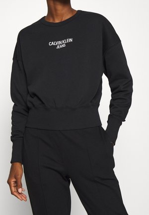 Osoba nosząca czarną bluzę Calvin Klein Jeans z prążkowanymi mankietami i paskiem, w połączeniu z czarnymi spodniami, na białym tle.