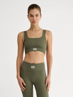 Vrouw staat en draagt een olijfgroen geribd sportbeha en bijpassende high-waist legging, beide met witte merklabels.