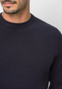 Marineblauer, gestrickter Pullover mit rundem Ausschnitt, der ein strukturiertes Muster und gerippte Bündchen aufweist, aus Schulterhöhe gezeigt.