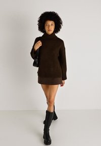 Brūns adīts kakla svārki ar ribotām malām, kopā ar īsu brūnu svārku un melniem garajiem zābakiem. Modelis tur melnu somu.