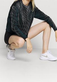 Czarny sportowy top z długimi rękawami z teksturowanym wzorem węża, w połączeniu z czarnymi szortami i białymi trampkami z srebrnym „swooshem”.