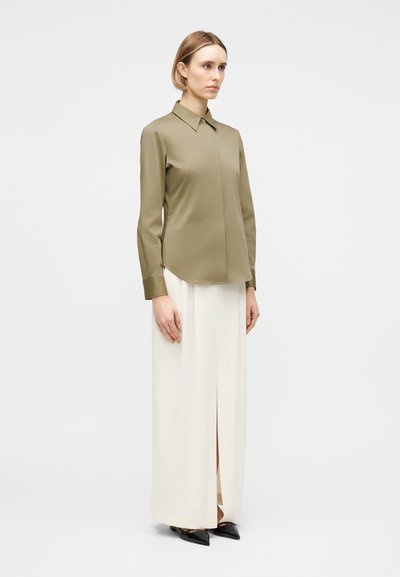 Γυναίκα όρθια, φορώντας πουκάμισο μακρυμάνικο ελιάς και ψηλόμεσο, λευκό παντελόνι φαρδύ με σκίσιμο μπροστά, συνδυασμένο με μαύρα μυτερά παπούτσια.