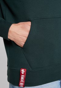 Felpa verde con tasca frontale, polsini e orlo a coste, caratterizzata da un'etichetta rossa con testo bianco sul lato inferiore. Realizzata in morbido cotone.