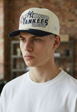 Young man wearing a white New York Yankees 1903 baseball cap and a plain white t-shirt, indoors with blurred background.