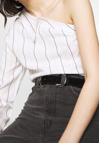 Camisa blanca a rayas con diseño de un solo hombro, combinada con jeans de talle alto de color gris oscuro y un cinturón negro con doble anilla.