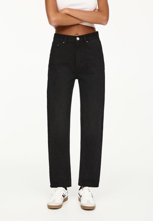 Person med armene over kors iført højtaljede sorte jeans med lige ben, hvid crop top og hvide sneakers med sorte striber.