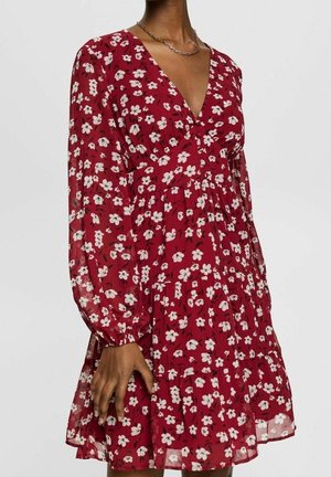 Vrouw draagt een lang mouw, rode jurk met bloemenprint, een V-hals en een getailleerde taille, gecombineerd met een zilveren ketting.