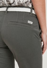 Pantalons en tissu gris avec une ceinture tissée blanche, présentant un détail de poche arrière et une étiquette "oxmo". Texture lisse, design ajusté.