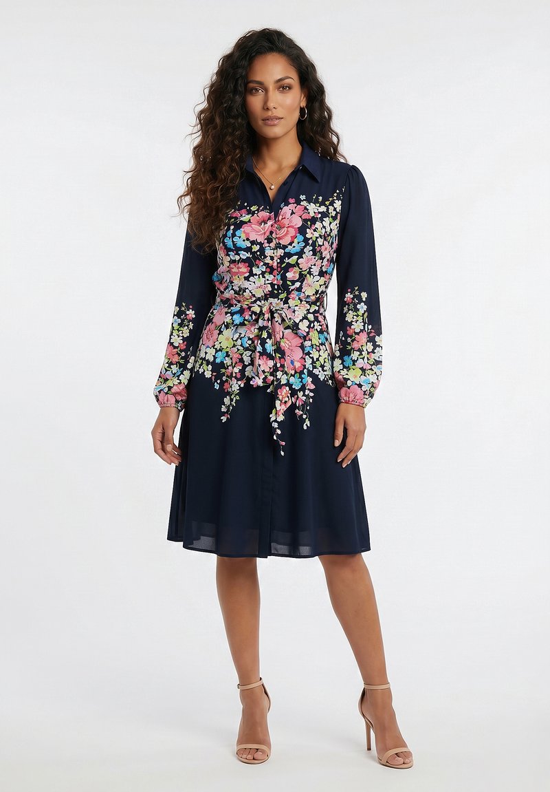 Femme aux longs cheveux bouclés portant une robe bleu marine à motifs floraux roses et bleus et des sandales à talons nus, debout sur un fond blanc.