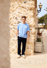 Blå bomullströja med en liten vit logotyp, tillsammans med mörka jeans och beige sneakers, framför en texturerad stenvägg.