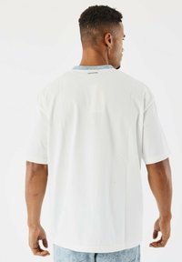 Hombre de espaldas con una camiseta blanca holgada y pantalones ligeros de color azul con estampado, contra un fondo blanco liso.