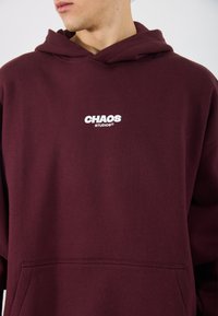 Hanorac mahon cu un logo alb "CHAOS STUDIOS" pe piept, având un buzunar frontal și un design clasic al glugii. Material din bumbac.