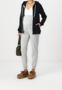 Sweat à capuche noir à fermeture éclair, joggeurs gris, haut blanc, chaussures à plateforme marron. Le mannequin tient un sac vert. Tissu lisse, coupe décontractée, couleurs contrastées.