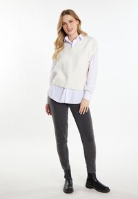 Maglione bianco senza maniche a maglia con scollo a V sopra una camicia azzurro chiaro con colletto, abbinato a pantaloni grigi e stivaletti neri alla caviglia.