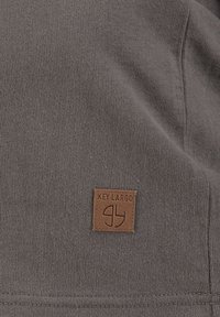 Dunkelgrauer Stoff mit glatter Textur verfügt über ein braunes Lederpatch, das mit "KEY LARGO" und einem kreisförmigen Design geprägt ist.