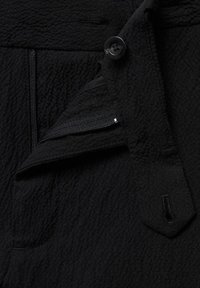 Tissu noir texturé présentant un bouton, une poche zippée et un petit rabat, mettant en valeur l'aspect doux et froissé du matériau.