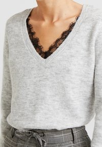 Grauer Pullover mit V-Ausschnitt und schwarzem Spitzenbesatz. Weicher Stoff, lange Ärmel, kombiniert mit karierten, hochtaillierten Hosen.