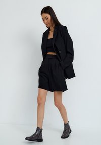 Zwarte blazer over een zwarte crop top en high-waisted shorts; model draagt glanzende zwarte enkelboots met veters en een gouden ketting.