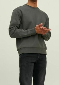 Sudadera de cuello redondo de un gris oscuro con puños y dobladillo acanalados, hecha de una tela suave, combinada con jeans oscuros, llevada por una persona con las manos entrelazadas.