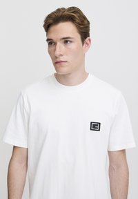 Hvit bomulls t-skjorte med korte ermer, med en liten svart brodert logo på venstre brystområde. Enkel og avslappet passform.