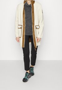 Giacca di pile crema e marrone con tasche frontali, abbinata a una maglietta scura a trama, pantaloni neri slim e sneakers nere con dettagli teal.