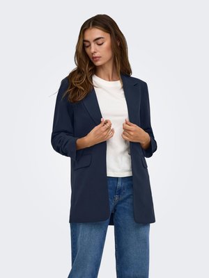Mujer con el cabello largo y castaño que lleva una blazer azul marino sobre una camiseta blanca y vaqueros azules, mirando hacia abajo y ajustándose las solapas de la blazer.