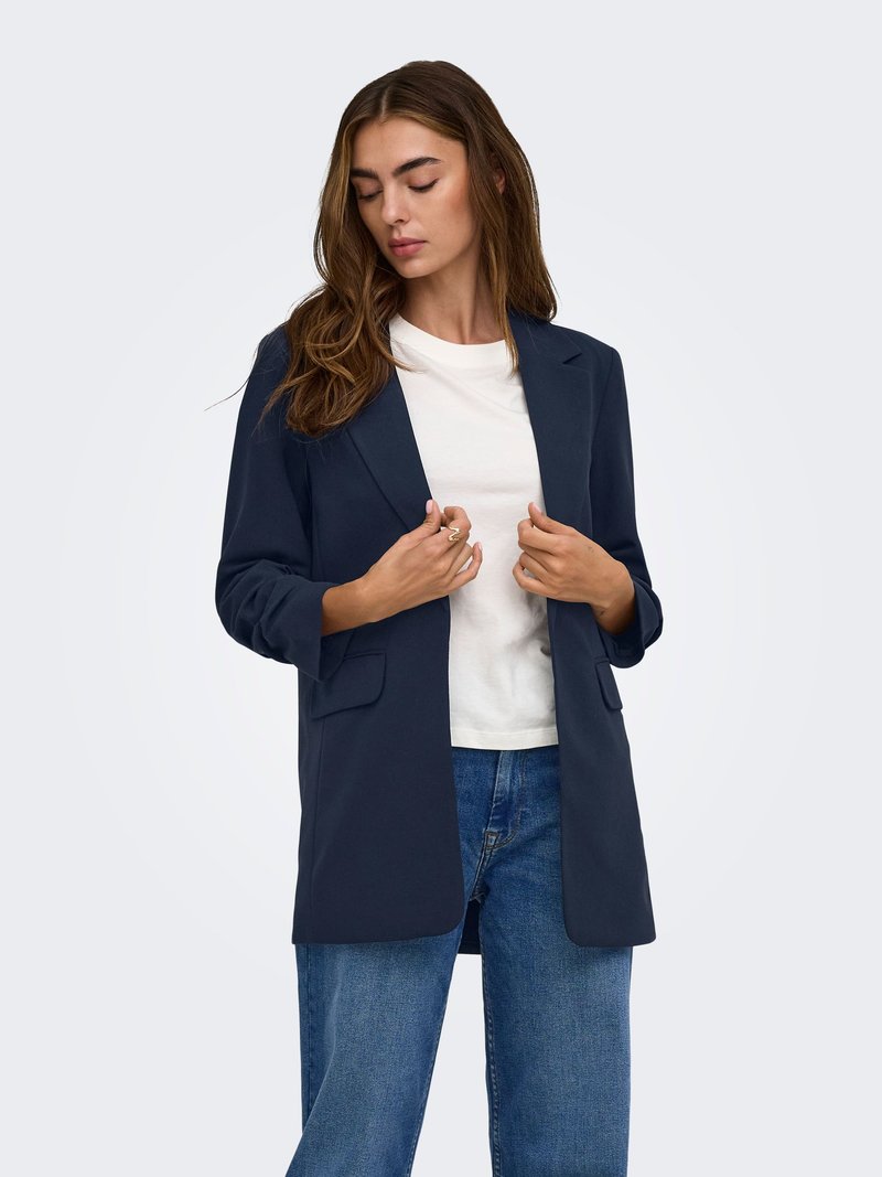 Femme aux longs cheveux bruns portant un blazer bleu marine par-dessus un t-shirt blanc et un jean bleu, regardant vers le bas et ajustant les revers du blazer.