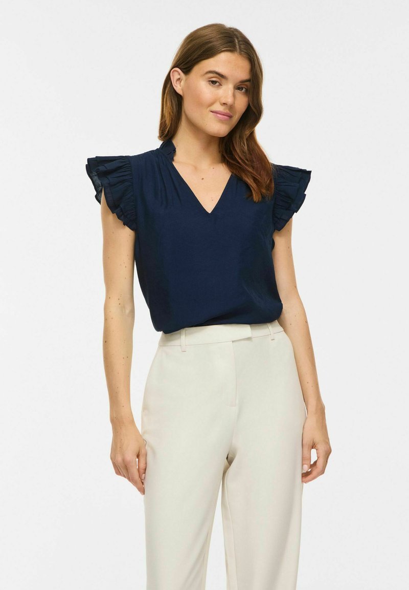 Femme portant un haut sans manches bleu marine avec des épaules volantées et un pantalon crème taille haute, debout devant un fond uni.
