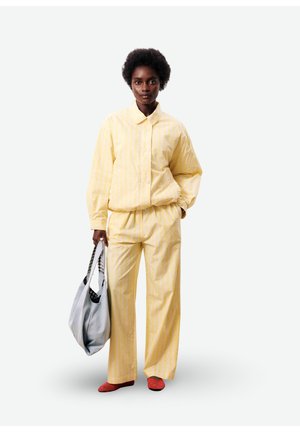 Femme portant une veste et un pantalon rayés jaune pâle, tenant un grand sac fourre-tout gris, debout devant un fond uni.