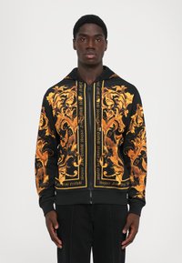 Haut à fermeture éclair noir avec des motifs floraux baroques dorés complexes et le texte "Versace Jeans Couture", porté par un homme debout.