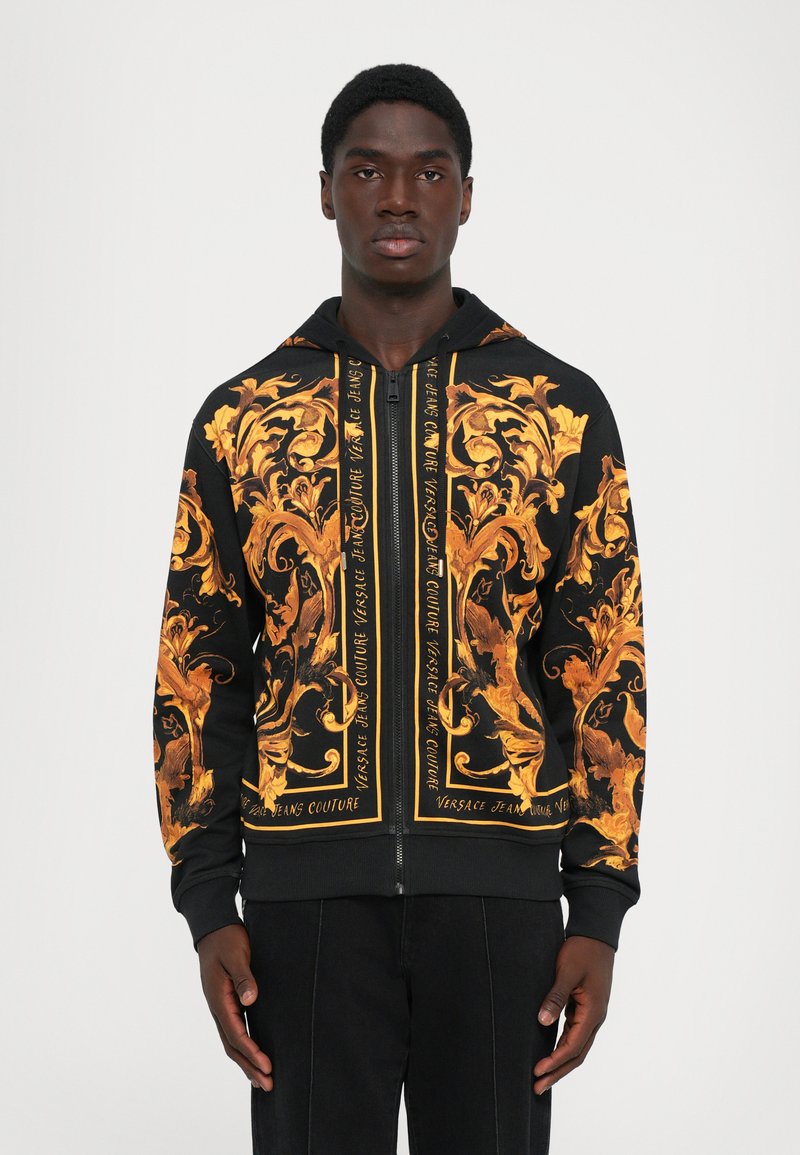 Haut à fermeture éclair noir avec des motifs floraux baroques dorés complexes et le texte "Versace Jeans Couture", porté par un homme debout.