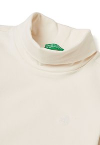 Krémový rolák s jemnou texturou, který má decentní vyšívaný emblém a zelenou etiketu s textem "United Colors of Benetton."