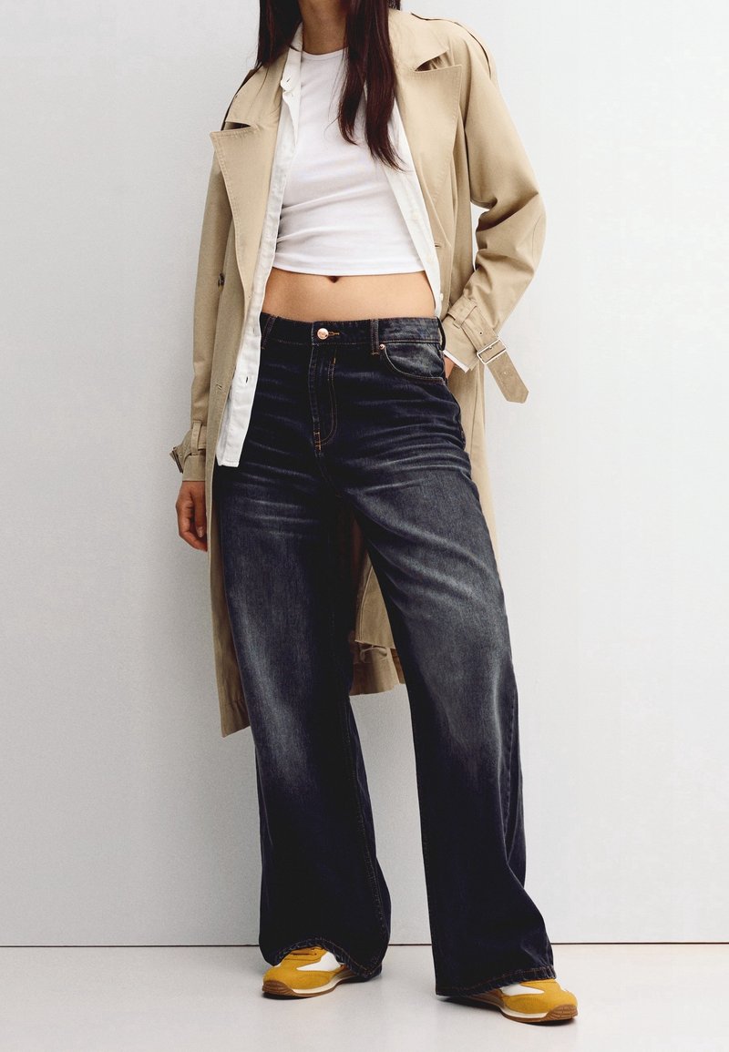 Femme portant un trench-coat beige, un crop top blanc, un jean large foncé et des baskets jaunes, debout contre un mur blanc uni.