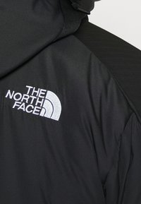 Veste noire à col haut avec un logo The North Face blanc brodé sur le côté gauche de la poitrine.