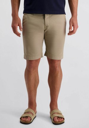 Mann trägt beigen knielangen Shorts und beigen Sandalen, steht vor einem einfarbigen hellen Hintergrund.