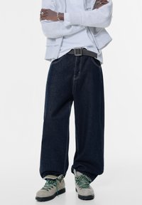 Lösa mörkblå denimjeans med synlig söm och midjeband med ett svart bälte, tillsammans med grå vandringsskor och gröna snören.