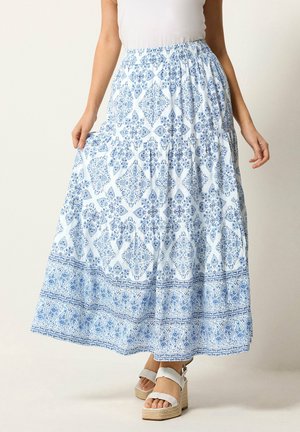 Mujer con un top blanco que lleva una falda larga con estampado floral blanco y azul, sosteniendo el lateral de la falda con la mano, y lleva sandalias blancas de plataforma.